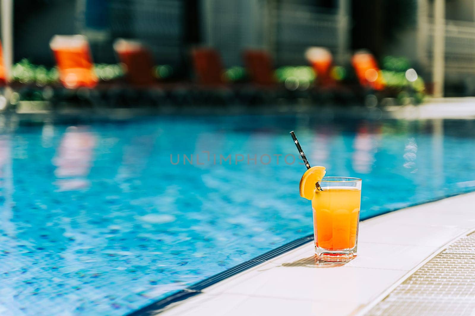 Tropical sparkling cocktail by the pool. The shot of glass with orange lemonade fruit cocktail standing near the poolside. Summer alcohol free drink by the hotel pool. Hello summer holiday vacation by Ostanina