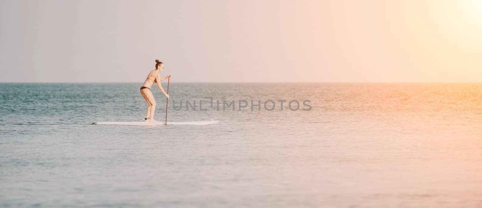 Silhouette of woman standing, surfing on SUP board, confident paddling through water surface. Idyllic sunset or sunrise. Sports active lifestyle at sea or river.