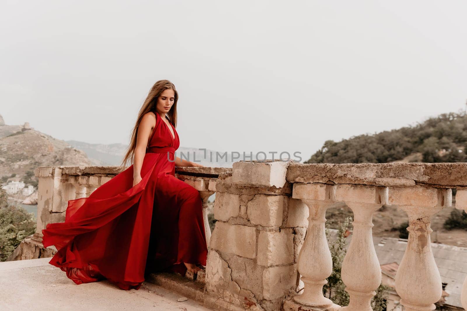 Side view a Young beautiful sensual woman in a red long dress posing on a volcanic rock high above the sea during sunset. Girl on the nature on overcast sky background. Fashion photo