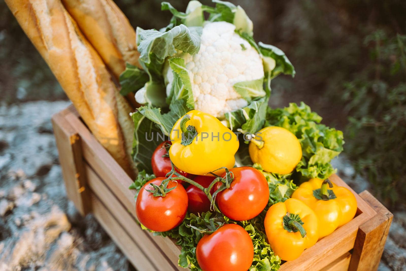 Wooden crate with farm market organic vegetables and baguettes standing on a stone. Closeup photo of frame crate with vegan food, tomatoes, bread, cauliflower, and sweet pepper