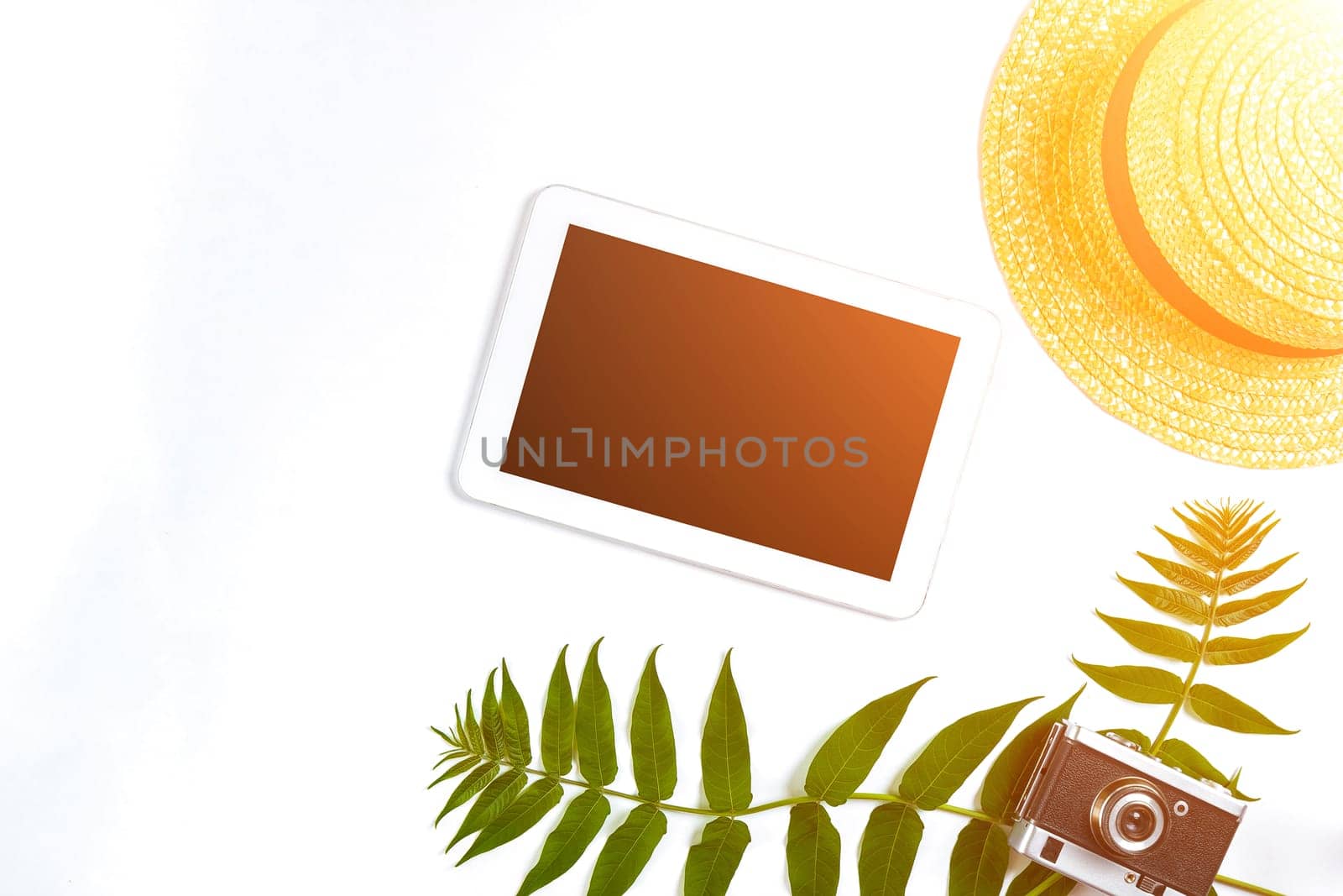 Straw hat with green leaves, tablet and old camera on white background, Summer background. Top view. Copy space. Still life. Flat lay. Sun flare