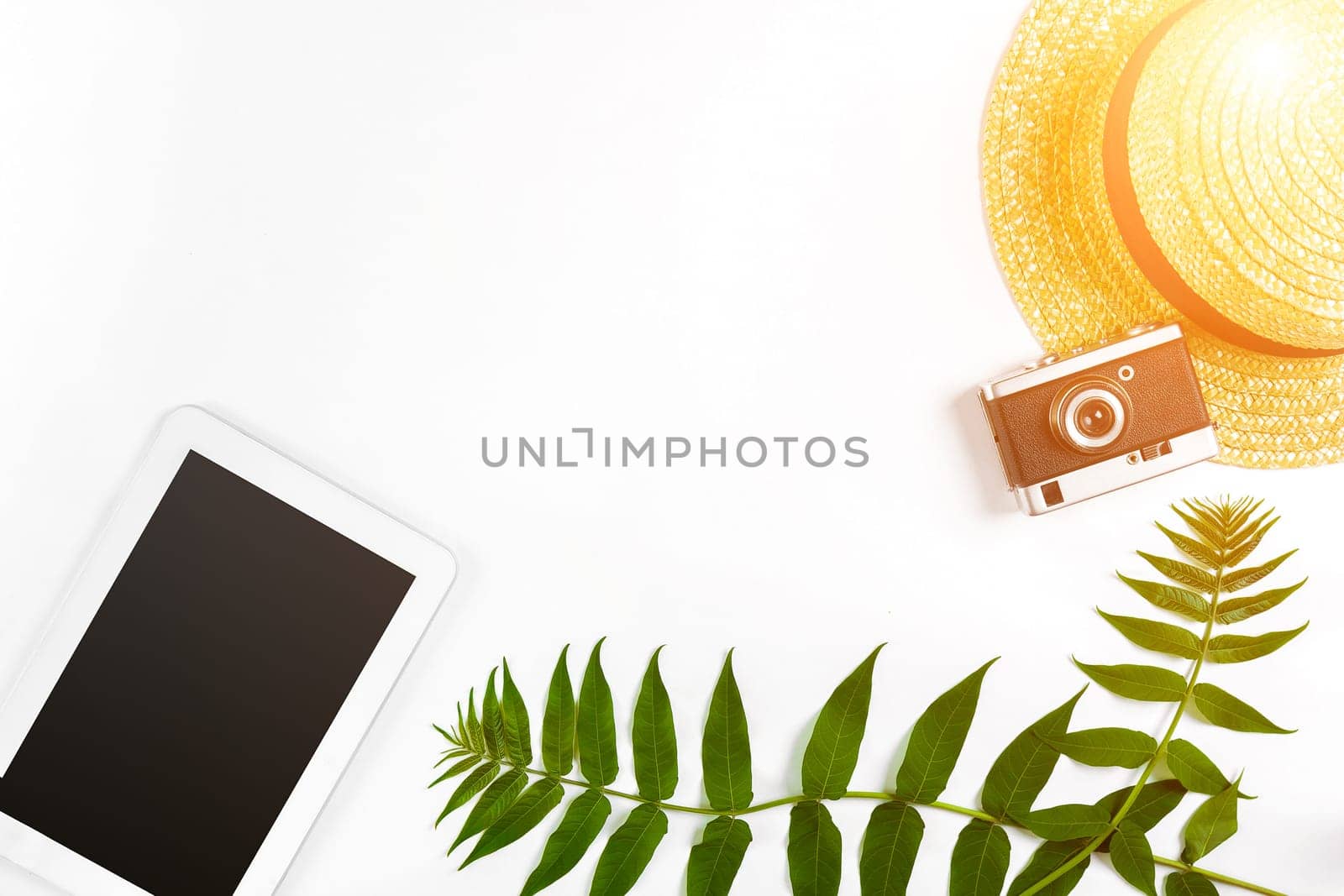 Straw hat with green leaves, tablet and old camera on white background, Summer background. Top view. Copy space. Still life. Flat lay. Sun flare