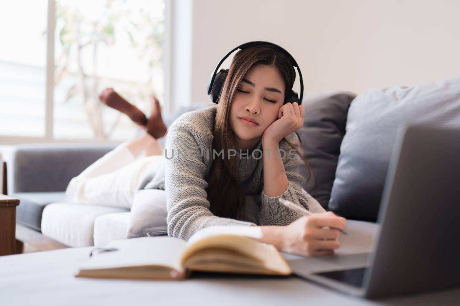 Online education, e-learning. Asian woman in stylish casual clothes, studying using a laptop, listening to online lecture, taking notes, online study at home by itchaznong