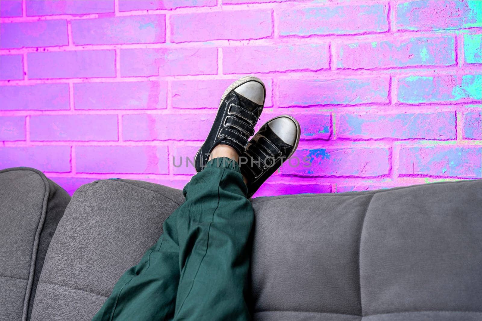 Relaxed legs of a boy in green casual pants and comfortable sneakers on a cozy sofa in the living room against a neon violet brick wall. children's feet in fashionable sneakers lie on the back of the sofa. Space for copying. Light violet neon brick background