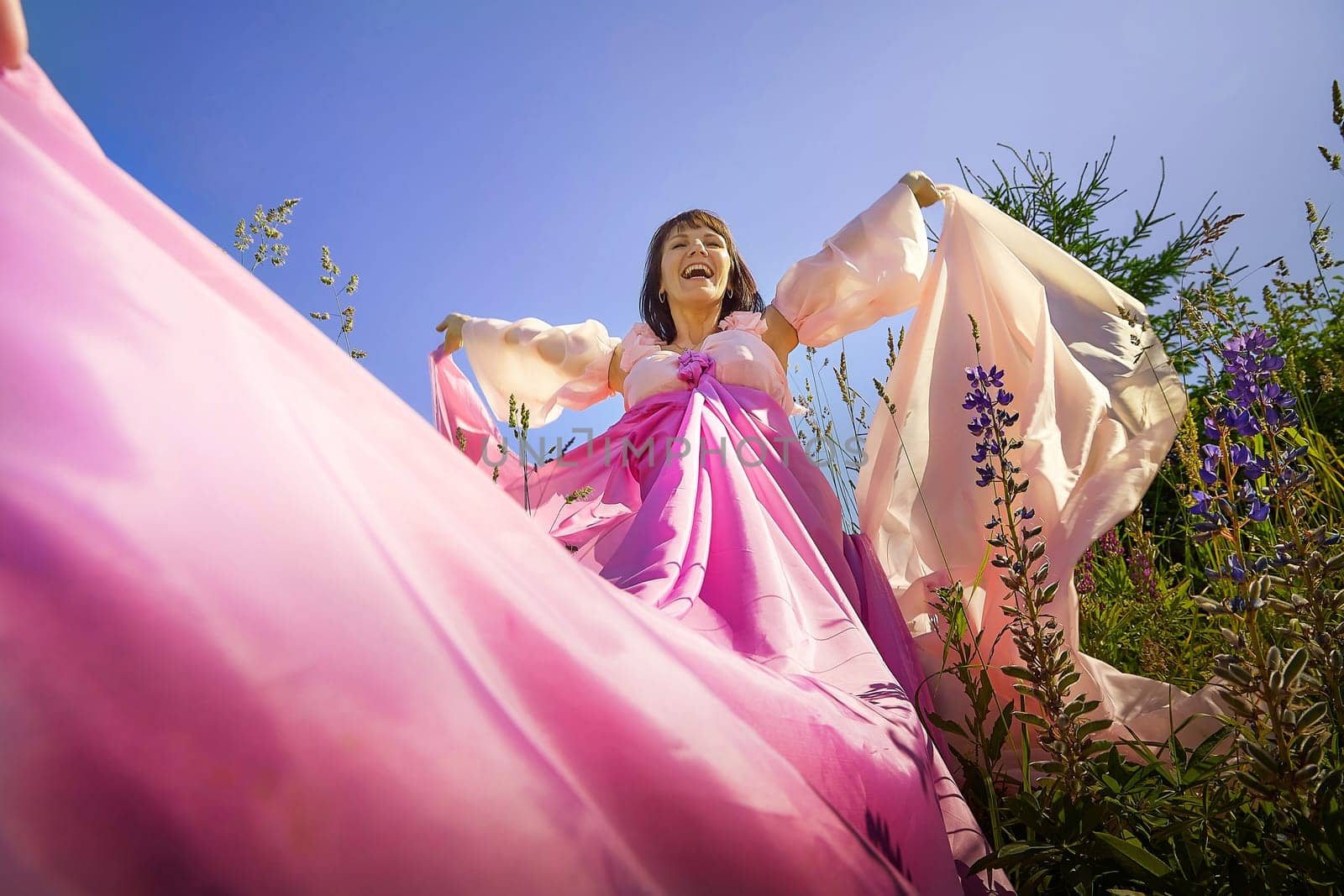 Beautiful girl in lush pink ball gown in green field during blooming of flowers and blue sky on background. Model posing on nature landscape as princess from fary tale