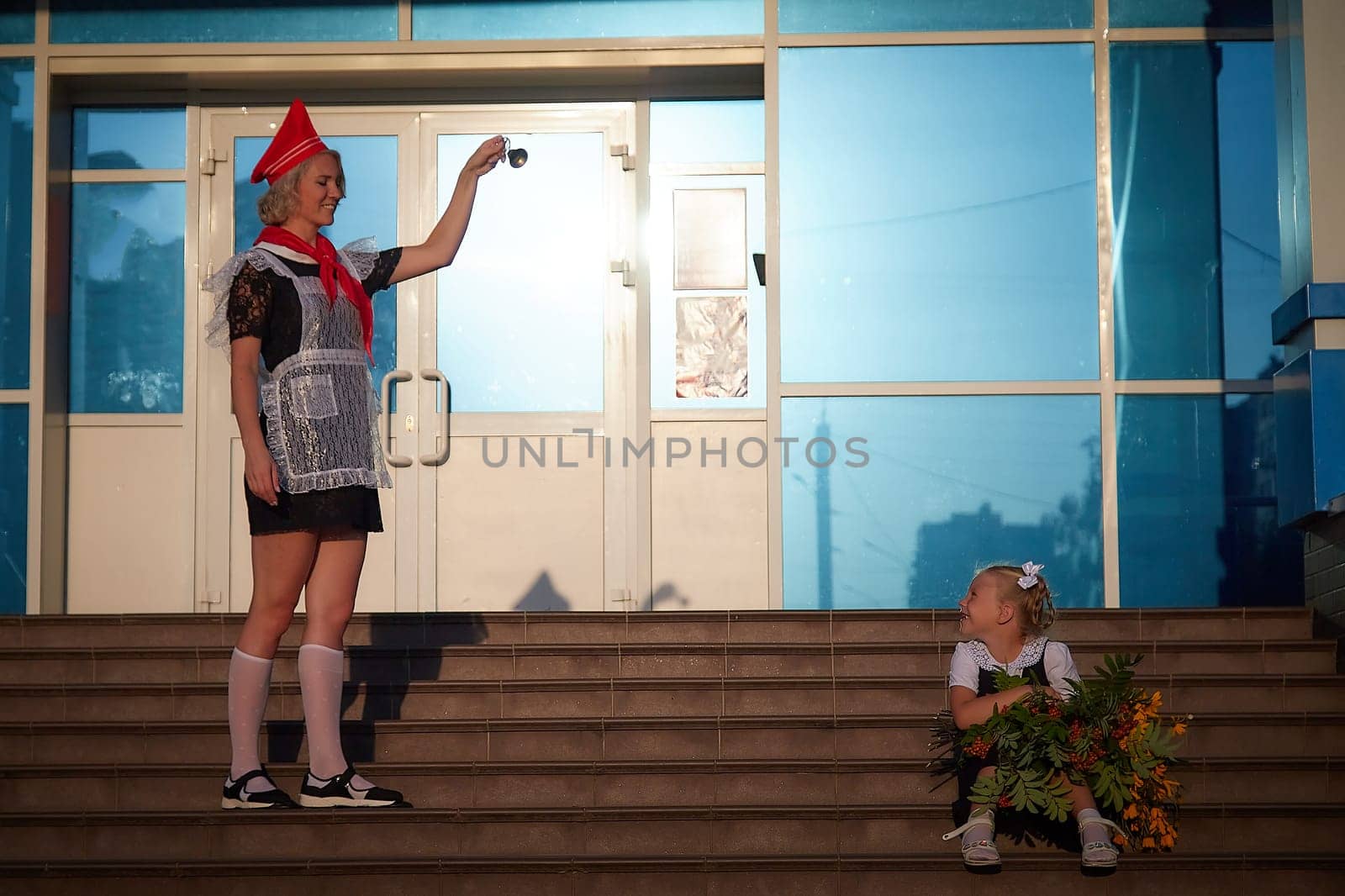 Young and adult schoolgirl on September 1 with flowers and bell. Generations of USSR and Russia. Female pioneer in red tie and October girl in modern uniform. Mother and daughter having fun by keleny