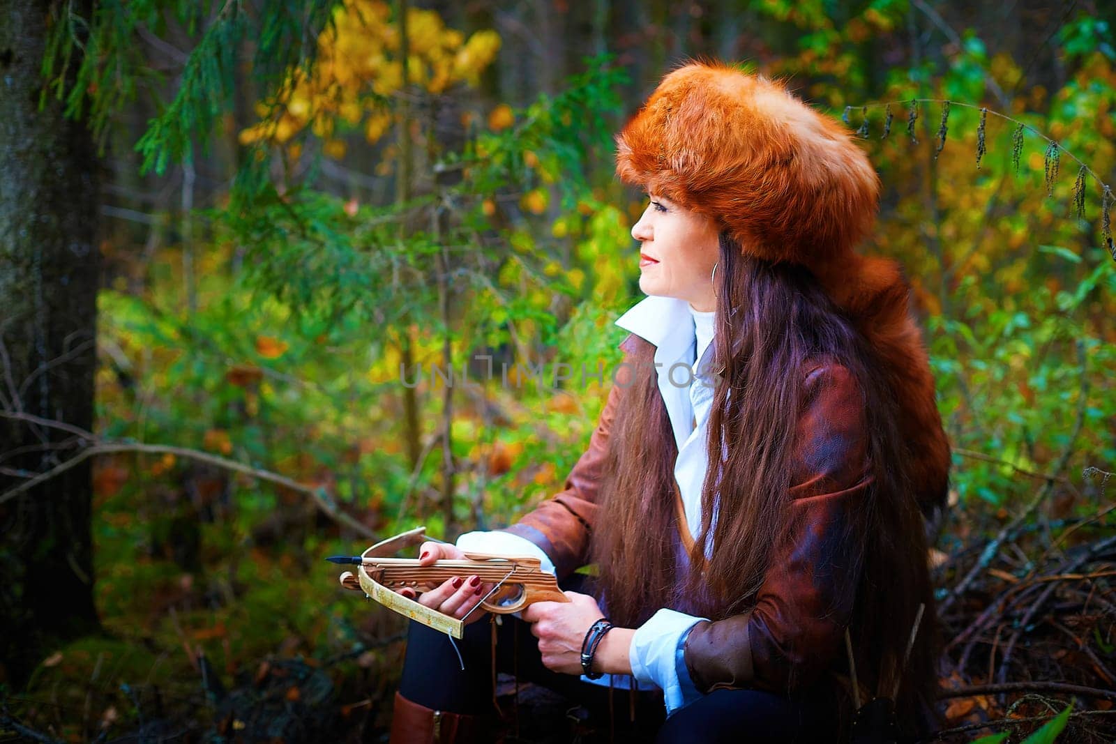 Girl in a leather jacket, a big red fox fur hat and with a crossbow in the forest in autumn. A female model poses as a fabulous royal huntress on a nature hunt at a photo shoot by keleny