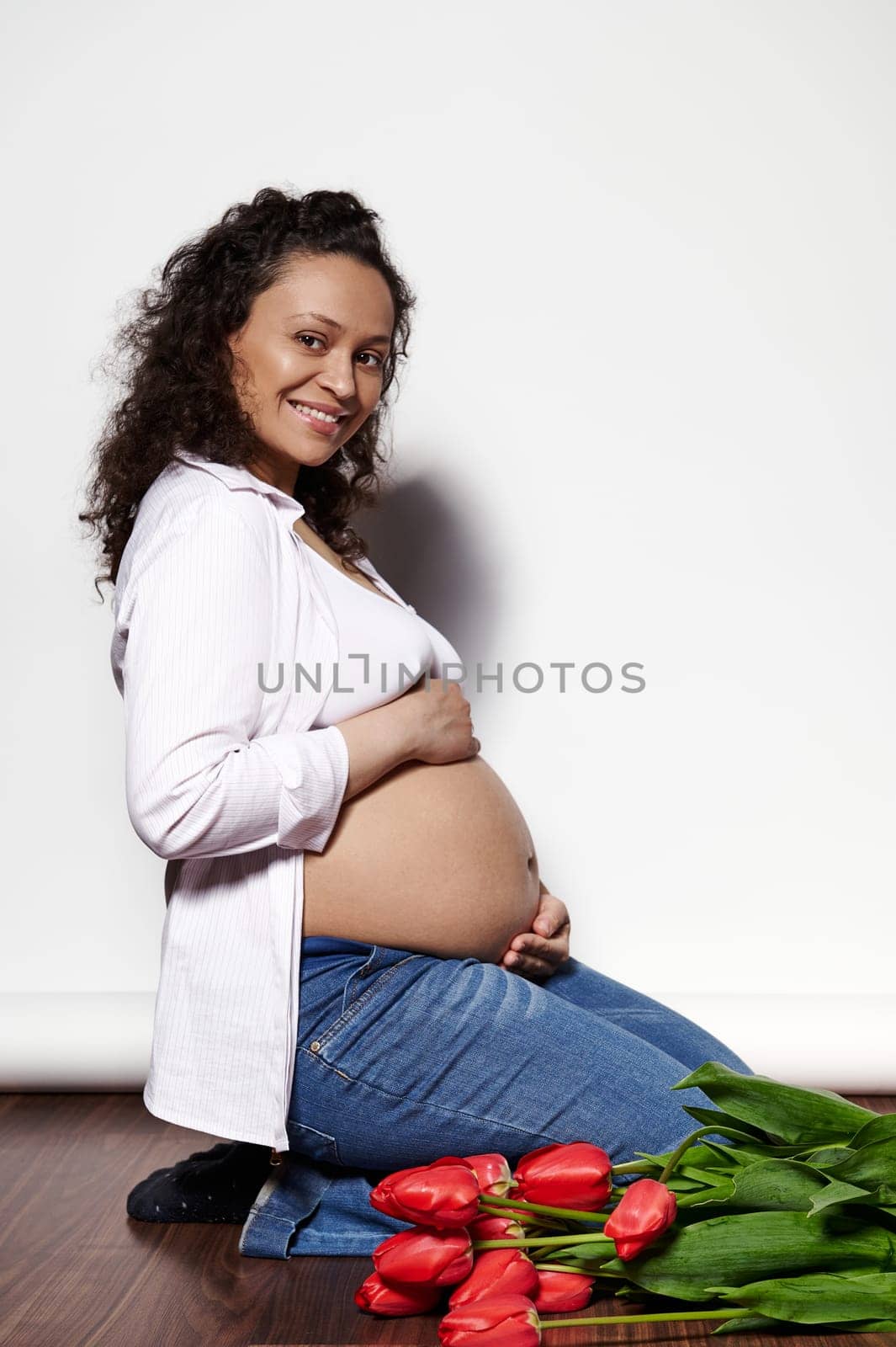 Ethnic curly happy adult pregnant woman, holding hands on belly, smiling looking at camera, isolated over white backdrop by artgf