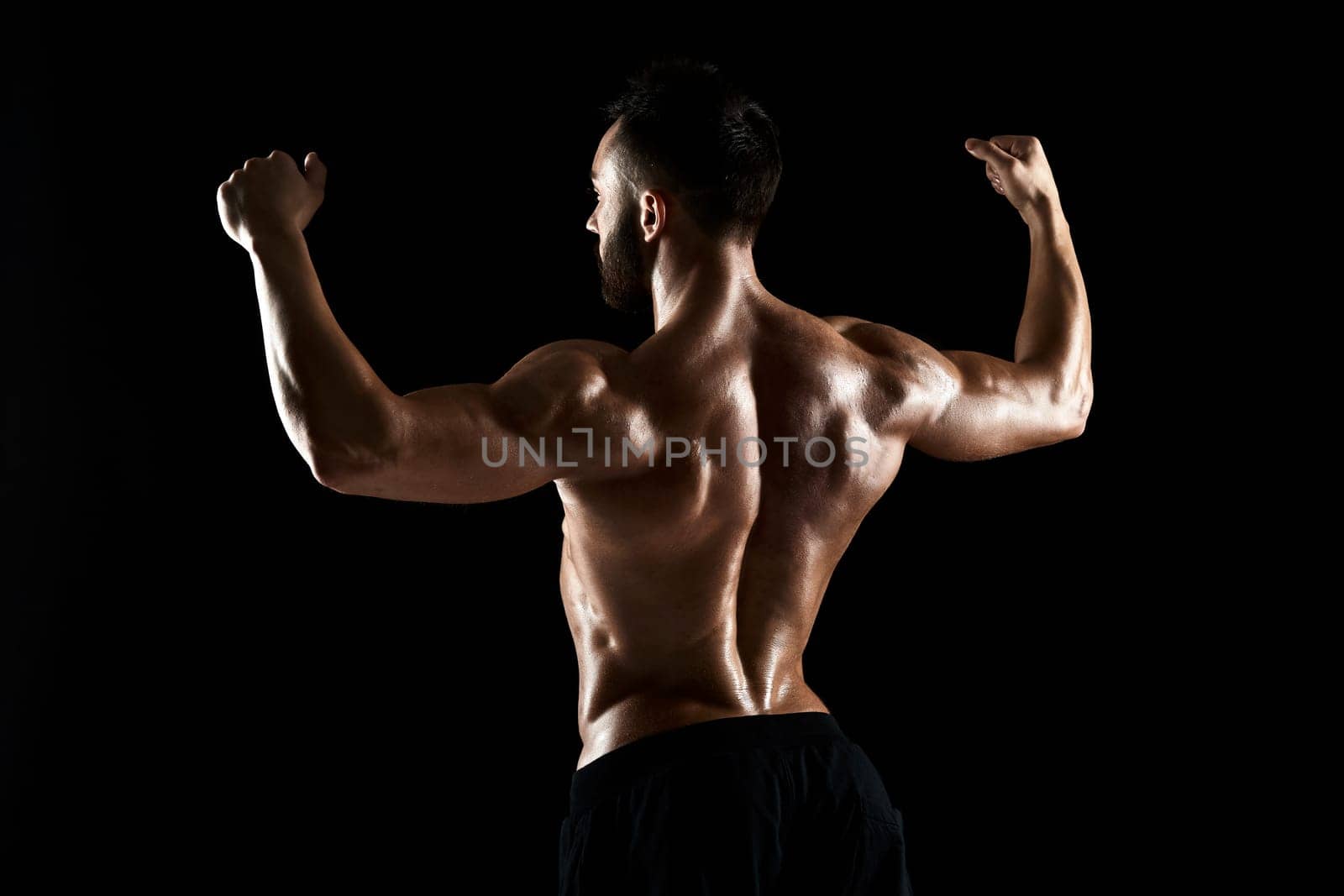 Image of very muscular man posing with naked torso in studio on black background. Rear view