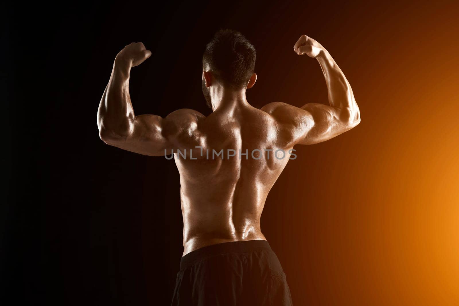 Image of very muscular man posing with naked torso in studio on black background. Rear view. with sun flare