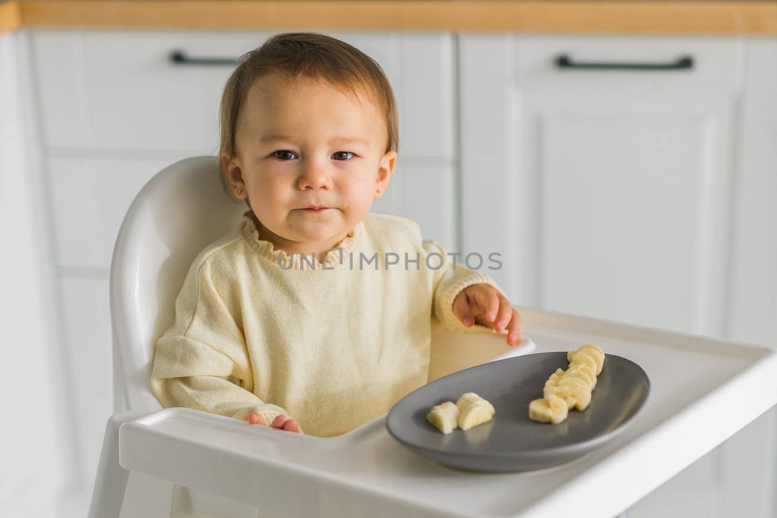 Happy baby sitting in high chair eating fruit in kitchen. Healthy nutrition for kids. Bio carrot as first solid food for infant. Children eat vegetables. Little boy biting raw vegetable. by Satura86