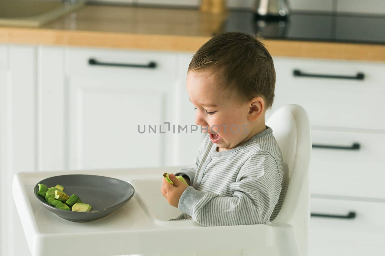 Happy baby sitting in high chair eating carrot in kitchen. Healthy nutrition for kids. Bio carrot as first solid food for infant. Children eat vegetables. Little boy biting raw vegetable