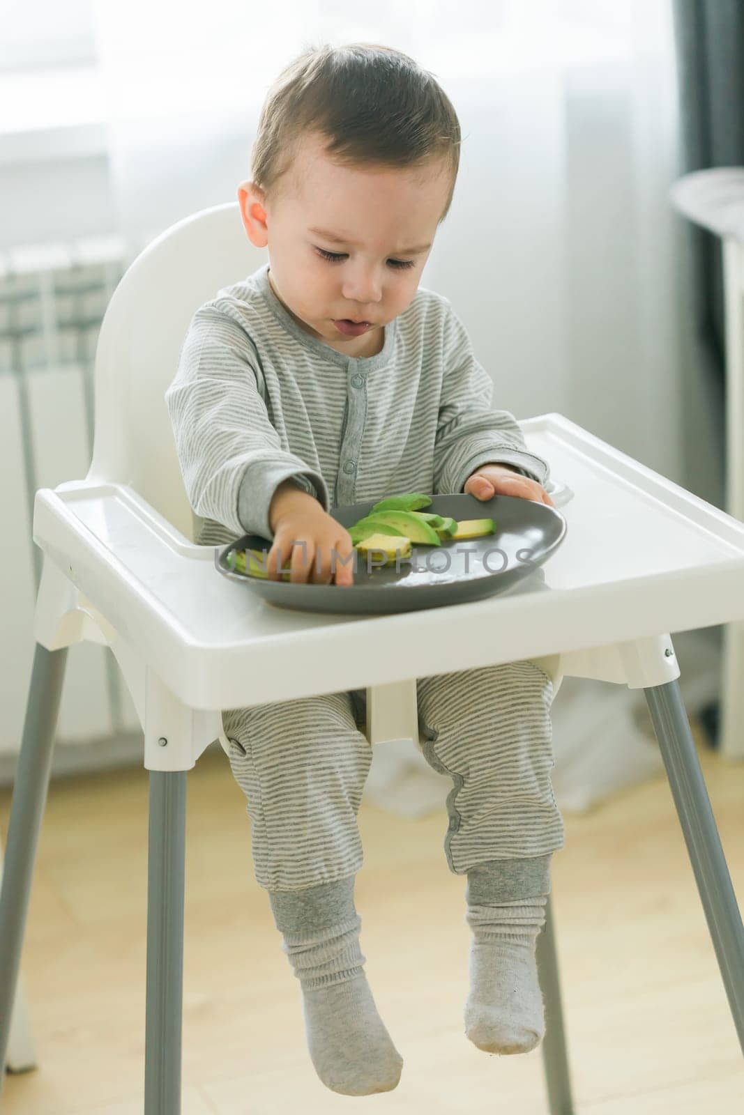 Happy baby sitting in high chair eating carrot in kitchen. Healthy nutrition for kids. Bio carrot as first solid food for infant. Children eat vegetables. Little boy biting raw vegetable