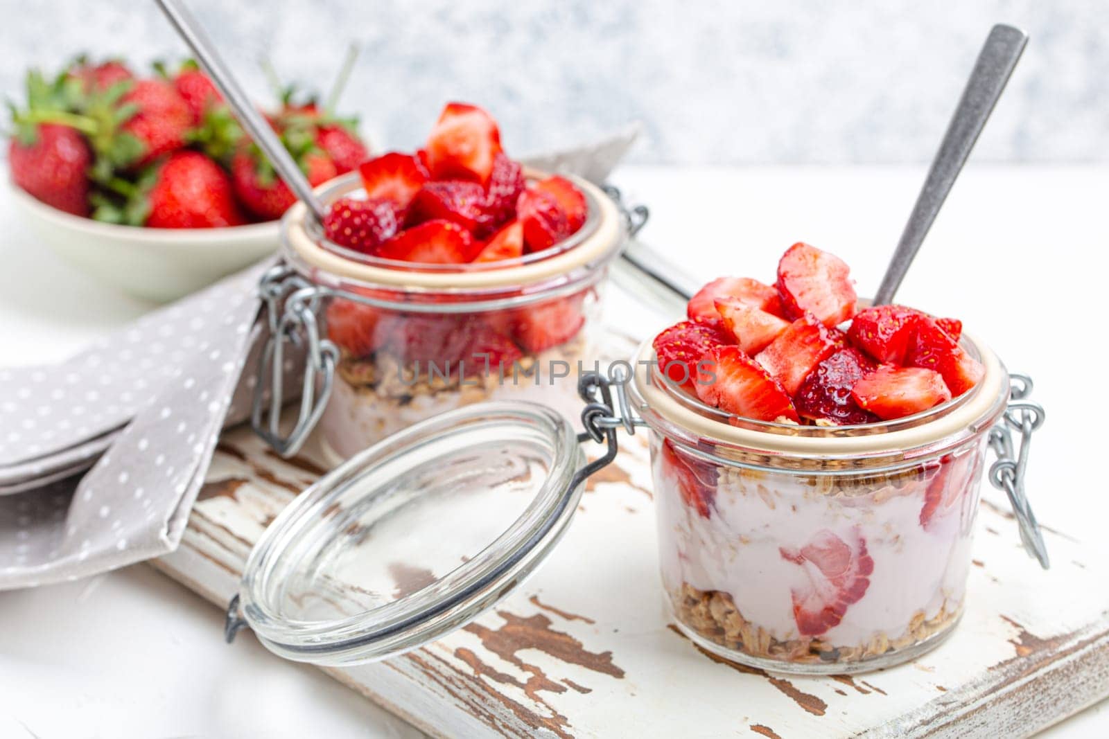 Parfait with Fresh Strawberries, Yoghurt and Crunchy Granola in Transparent Glass Mason Jars on White Rustic Wooden Background from Angle View, Healthy Breakfast or Light Summer Fruit Dessert