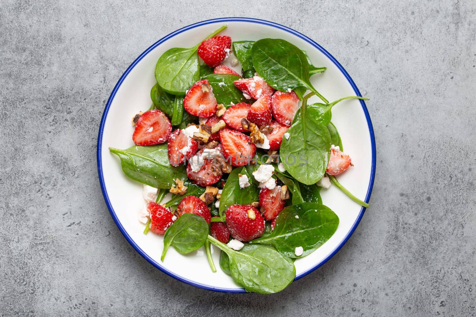 Light Healthy Summer Salad with fresh Strawberries, Spinach, Cream Cheese and Walnuts on White Ceramic Plate, Grey rustic stone Background Top View.