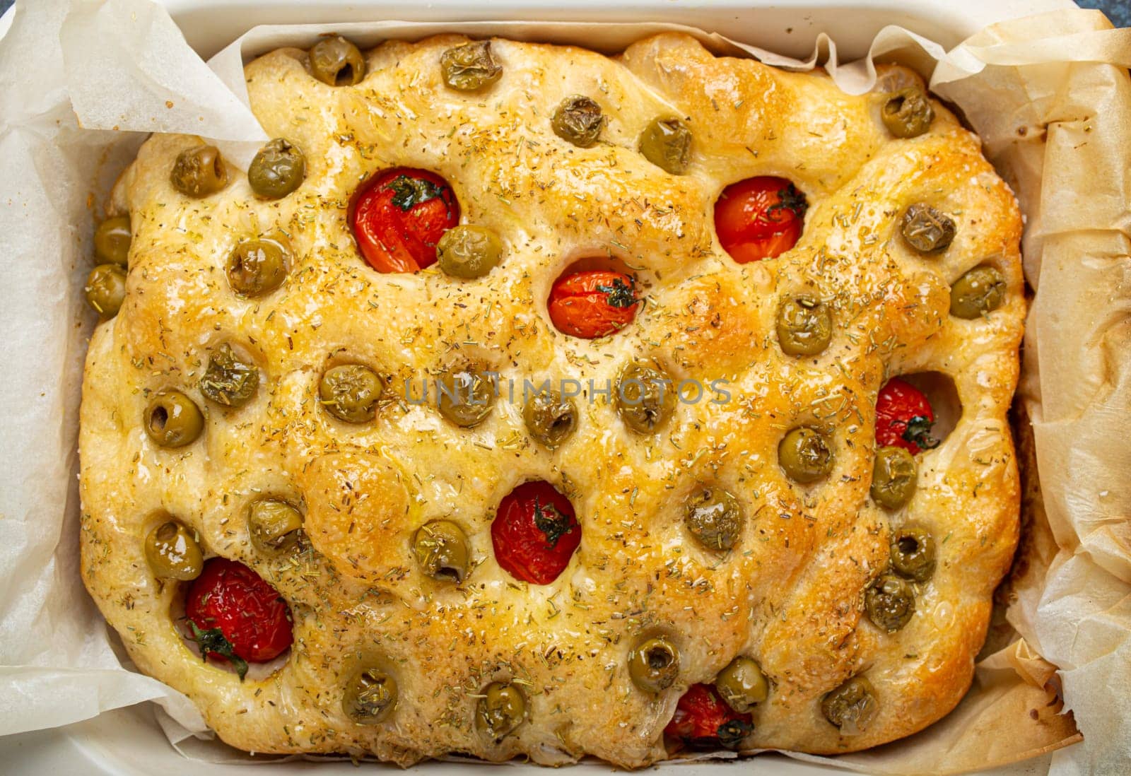 Overhead of Traditional Italian Homemade Flat Bread Focaccia with Green Olives, Olive Oil, Cherry Tomatoes and Rosemary in Baking Tray on Rustic Dark Blue Concrete Background