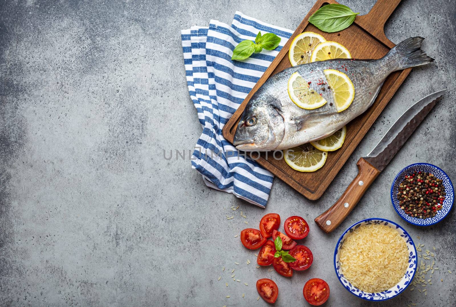 Raw fish dorado with ingredients lemon, fresh basil, cut cherry tomatoes, uncooked rice on wooden cutting board with knife on rustic stone background top view, cooking healthy fish dorado. Copy space