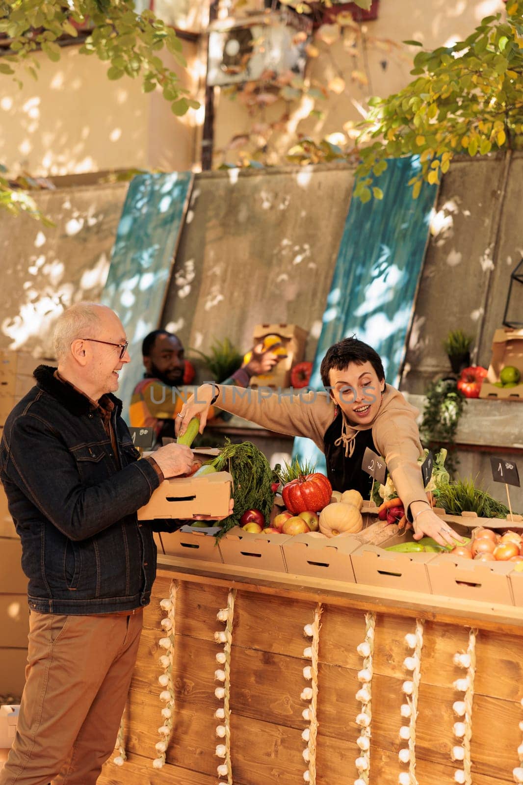 Smiling local farmer selling fresh bio natural products, giving food market fruits and vegetables to old customer. Female vendor talking to senior man about seasonal various healthy produce.