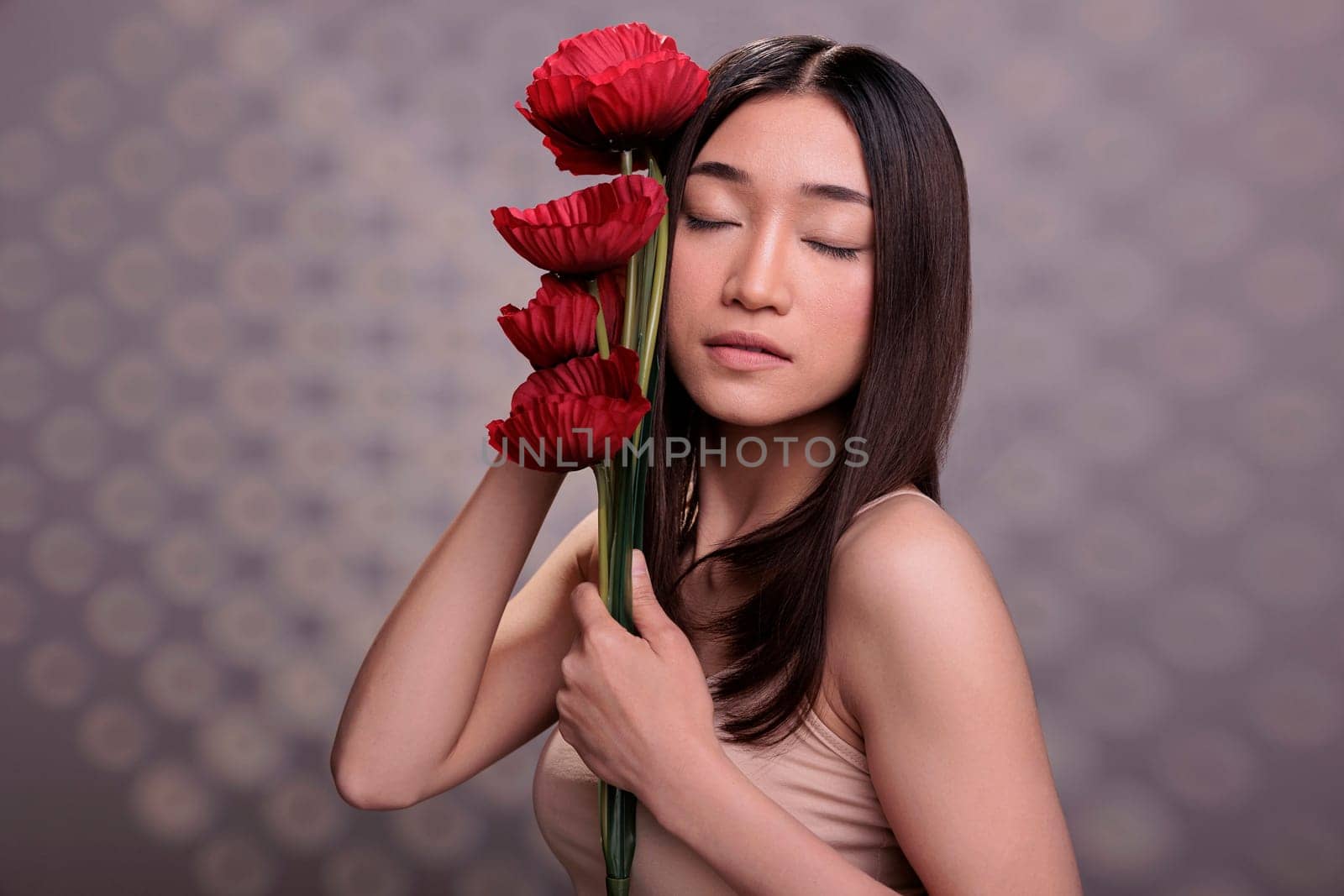 Asian woman with closed eyes holding red poppies branch. Beautiful young model wearing beige casual top, standing with summer flowers bouquet, showing skincare and body care concept