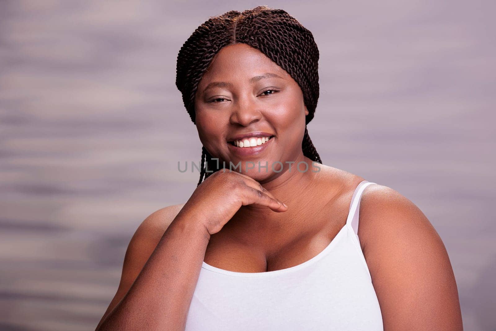 Attractive smiling african american curvy young woman holding hand on face portrait. Cheerful body positive lady putting arm on cheek, looking at camera and posing in studio