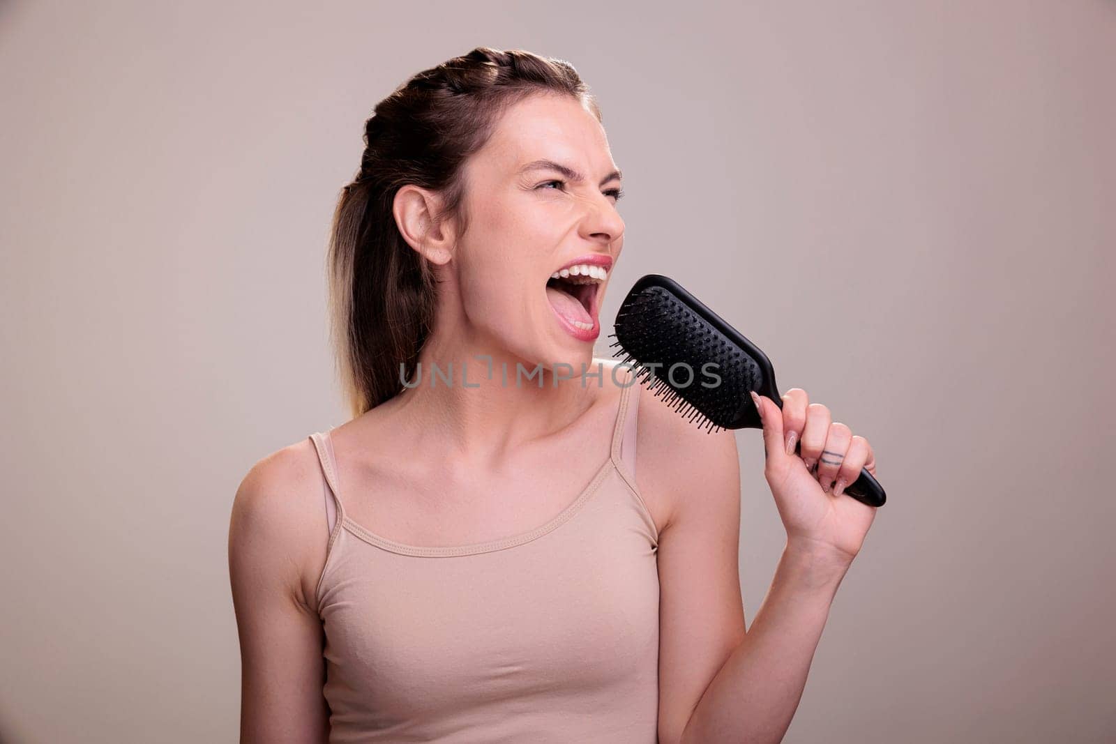 Beautiful woman singing loudly in hairbrush during hairstyle routine. Attractive young caucasian model posing with comb, using hairdressing tool as microphone and having fun