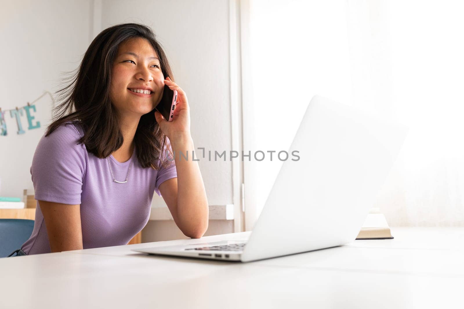 Happy Asian woman smiling talking on the phone while working at home office. College student using mobile phone. by Hoverstock