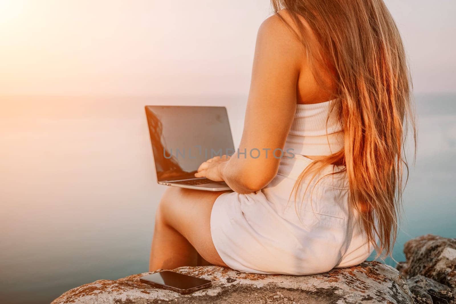 Woman laptop sea. Working remotely on seashore. Happy successful woman female freelancer in straw hat working on laptop by the sea at sunset. Freelance, remote work on vacation by panophotograph