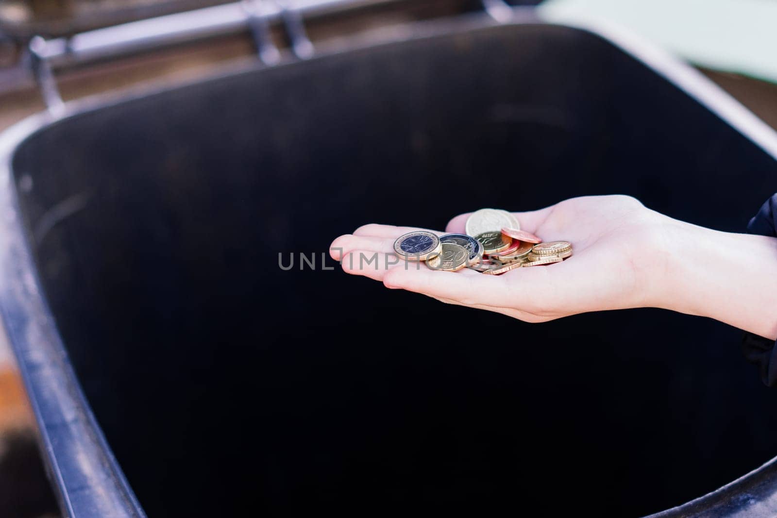 A kid girl hand throws euro coins into a trash can dumpster, street shot.