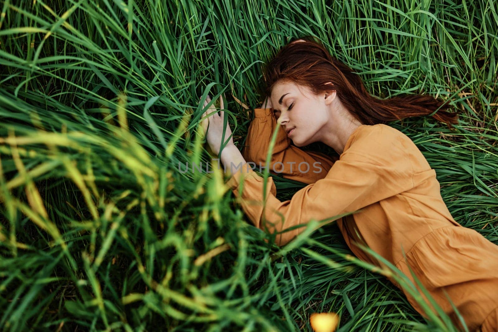 a close horizontal photo of a pleasant woman in a long orange dress resting lying in the tall grass with her eyes closed in sunny weather at sunset with her arms outstretched. Street photography, the theme of privacy with nature. High quality photo