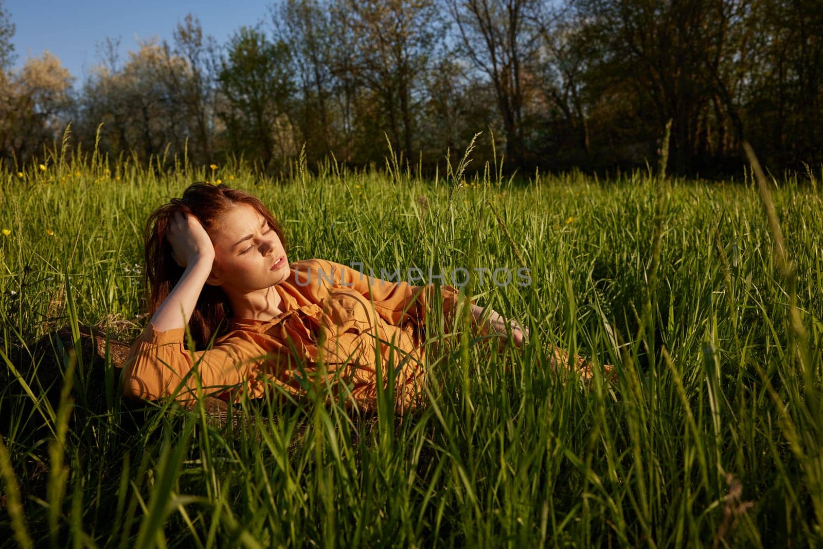 a beautiful, stylish woman lies in a long orange dress in the tall green grass illuminated by the sun rays with her eyes closed, holding her hand near her head by Vichizh