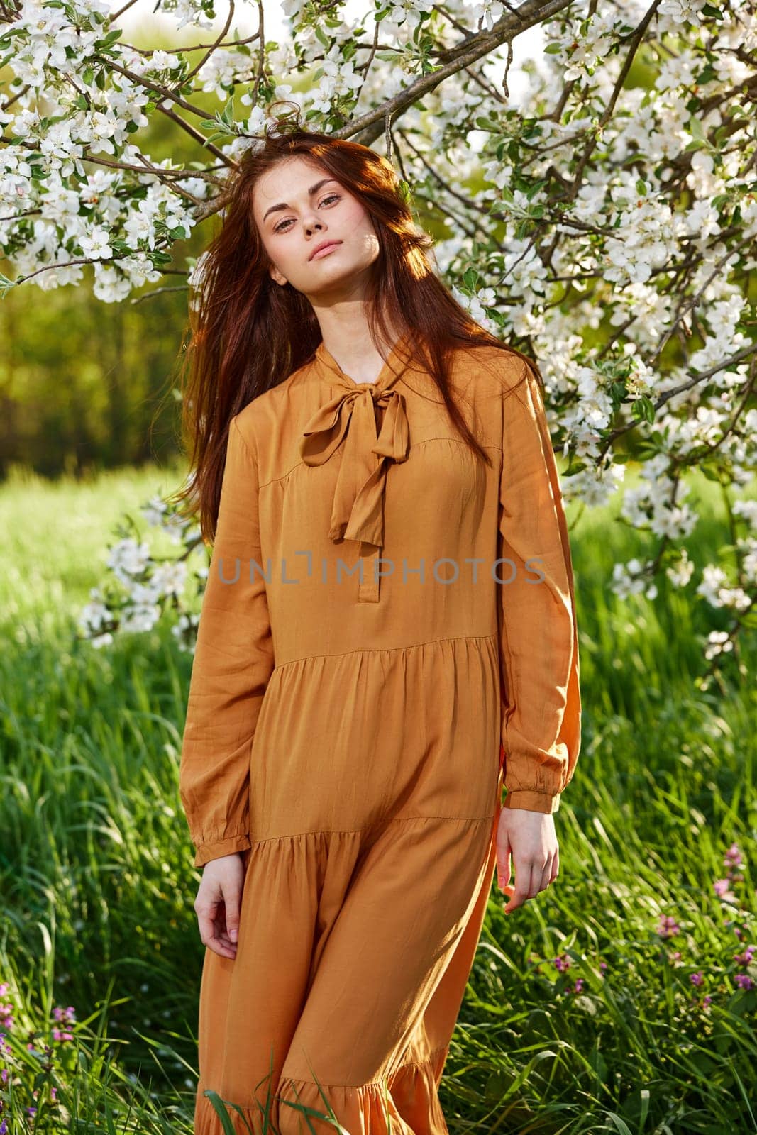 a bright vertical photo of an attractive woman in a long orange dress standing next to a flowering tree in sunny, warm weather, looking pleasantly into the camera. High quality photo