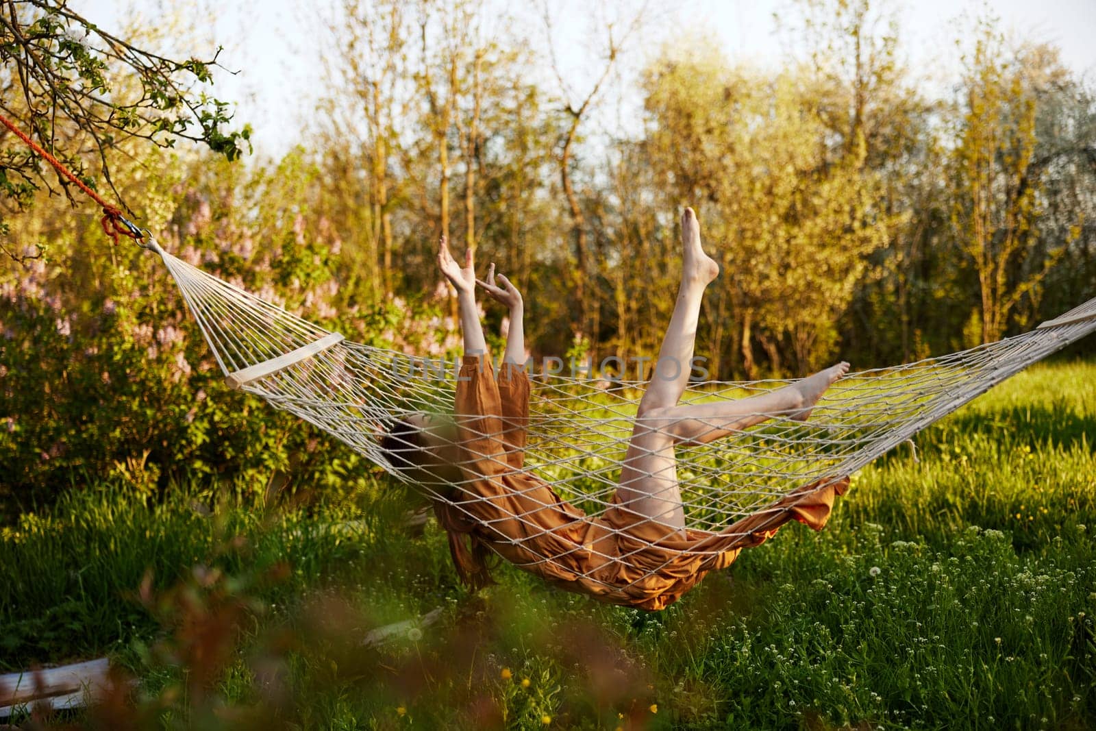 a funny woman is resting in nature lying in a mesh hammock in a long orange dress lifting up her arms and legs. High quality photo