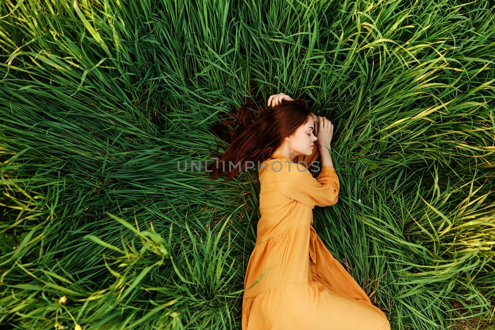 an elegant woman lies in the tall green grass in a long summer orange dress and holding her hair with her hands enjoys nature and sunny weather with her eyes closed by Vichizh