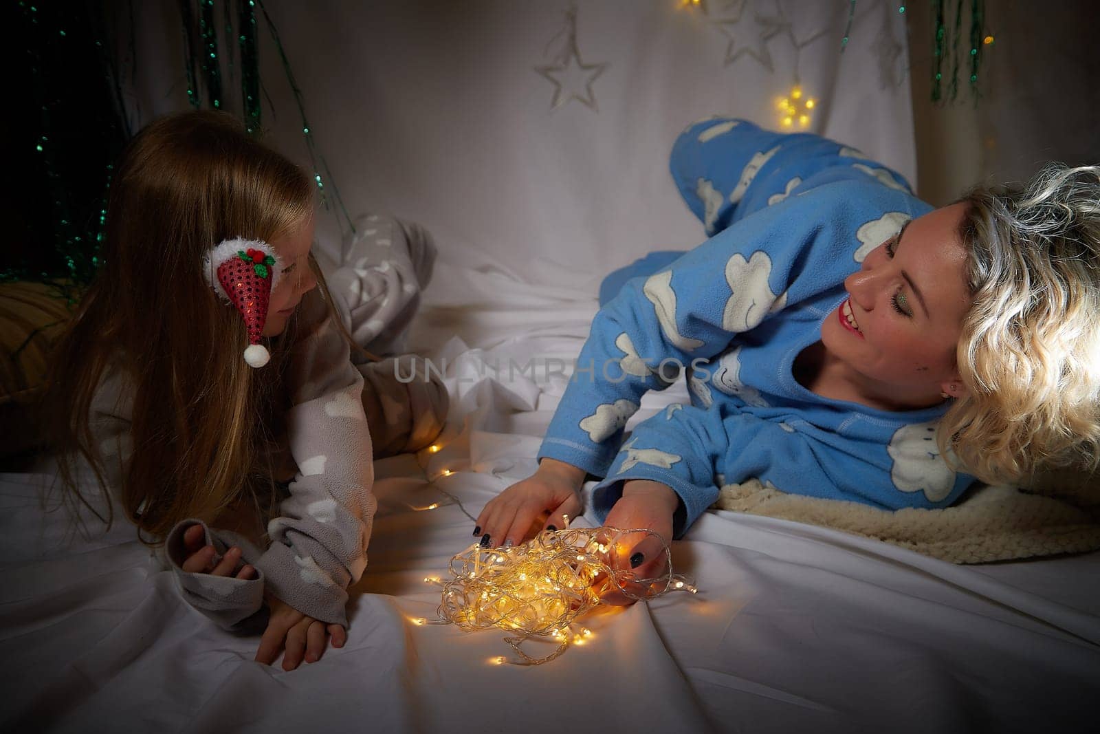 Cute mother and daughter in pajamas lie on soft blanket and having fun in room with Christmas garlands and white background. Tradition of decorating house for holiday. Happy childhood and motherhood