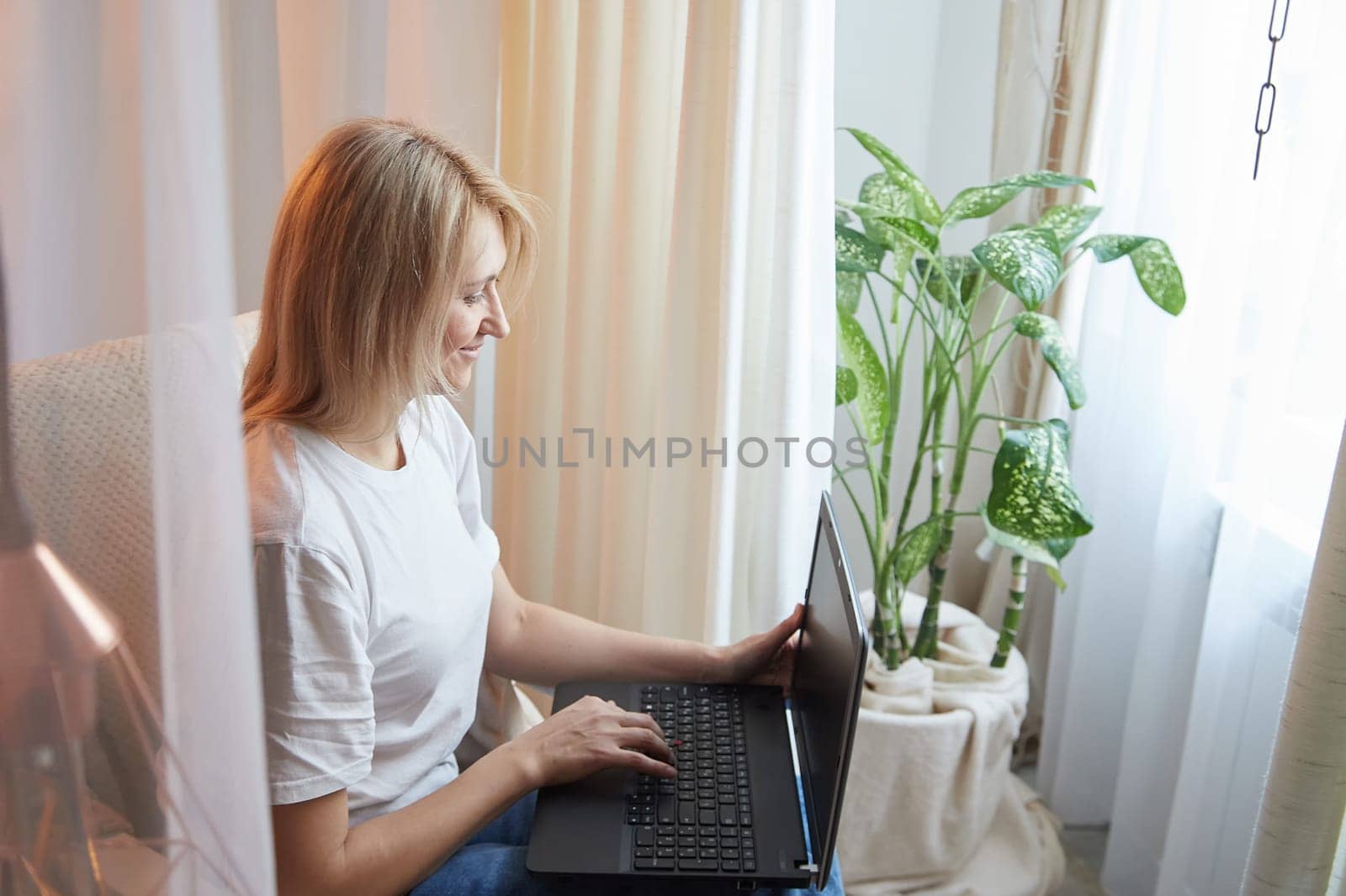 Young blonde woman using laptop computer at home. Girl is resting, chating and looking in camera near window at home. Nice atmosphere. The concept of cozy home