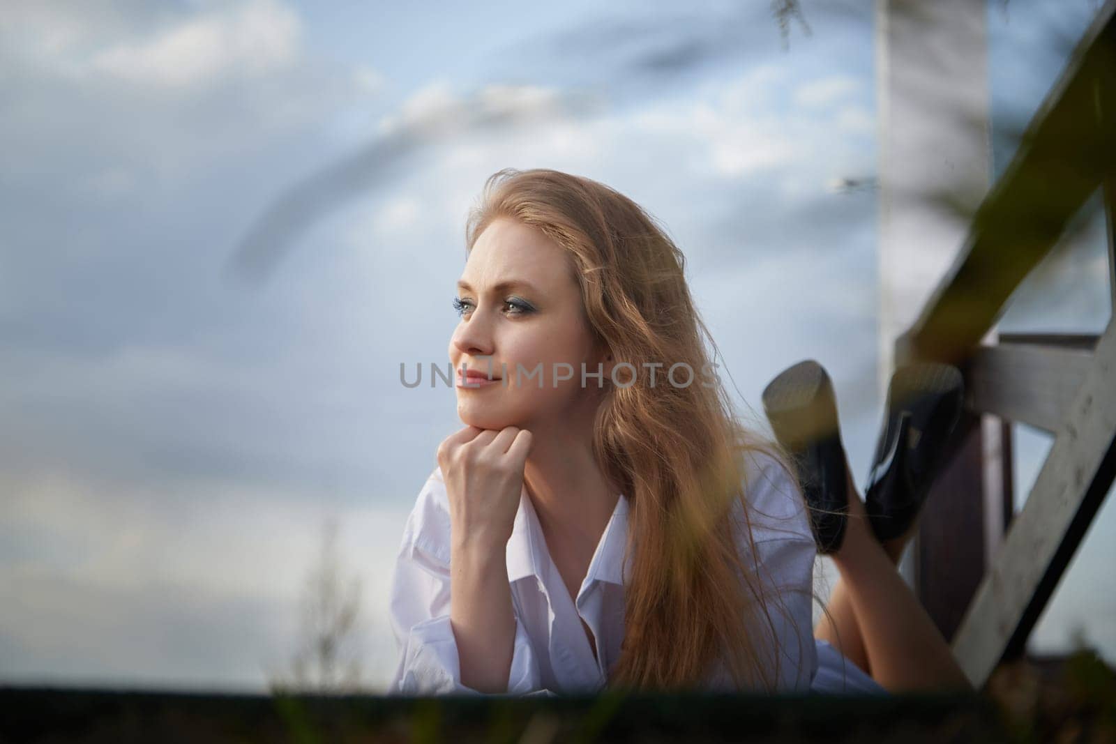 Beautiful girl with long hair in white shirt in open wooden pavillion in a village or small town. Young slender woman and sky with clouds on background on an autumn, spring or summer day
