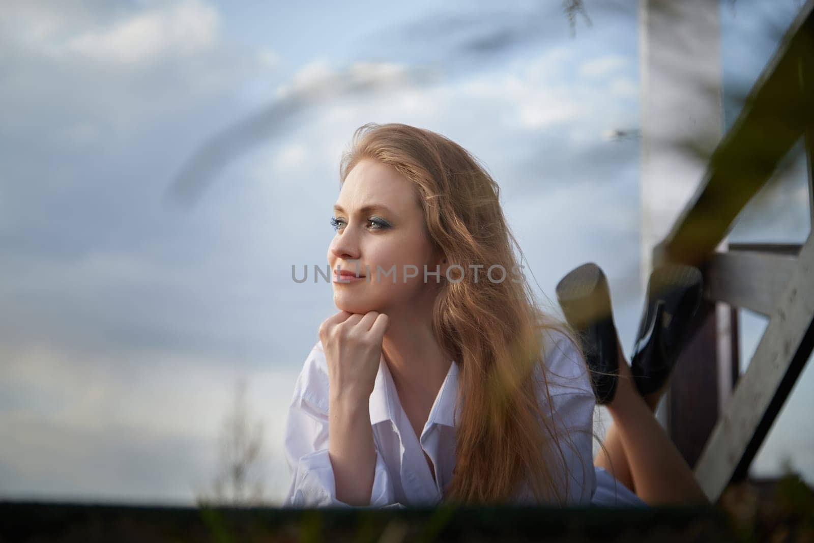 Beautiful girl with long hair in white shirt in open wooden pavillion in a village or small town. Young slender woman and sky with clouds on background on an autumn, spring or summer day
