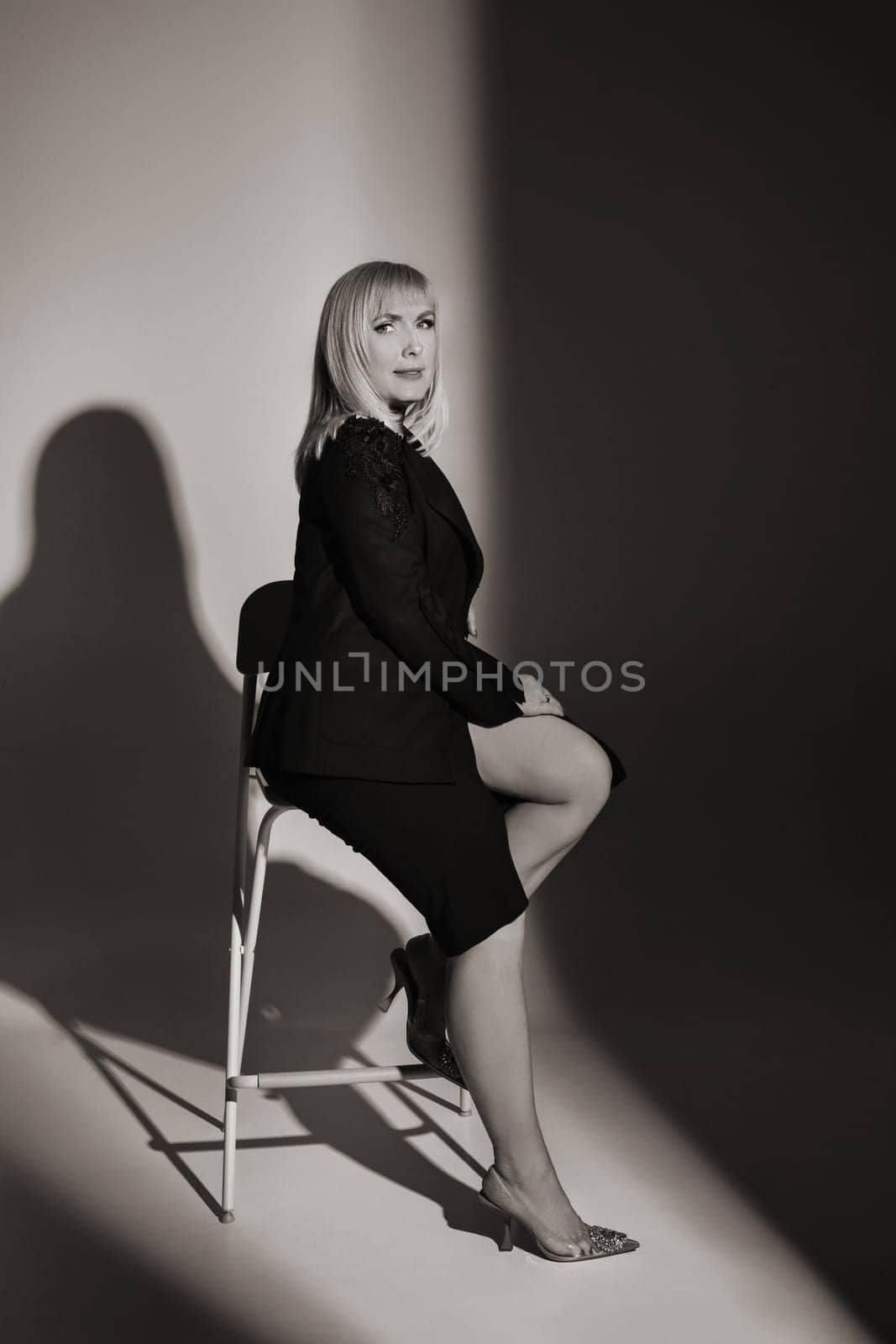 A fashionable woman in a black jacket and dress poses in a studio on a chair.
