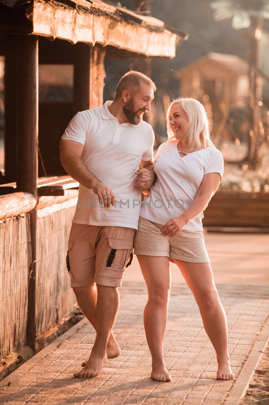 A happy married couple in shorts and T-shirts walks down the street at sunset in summer by Lobachad