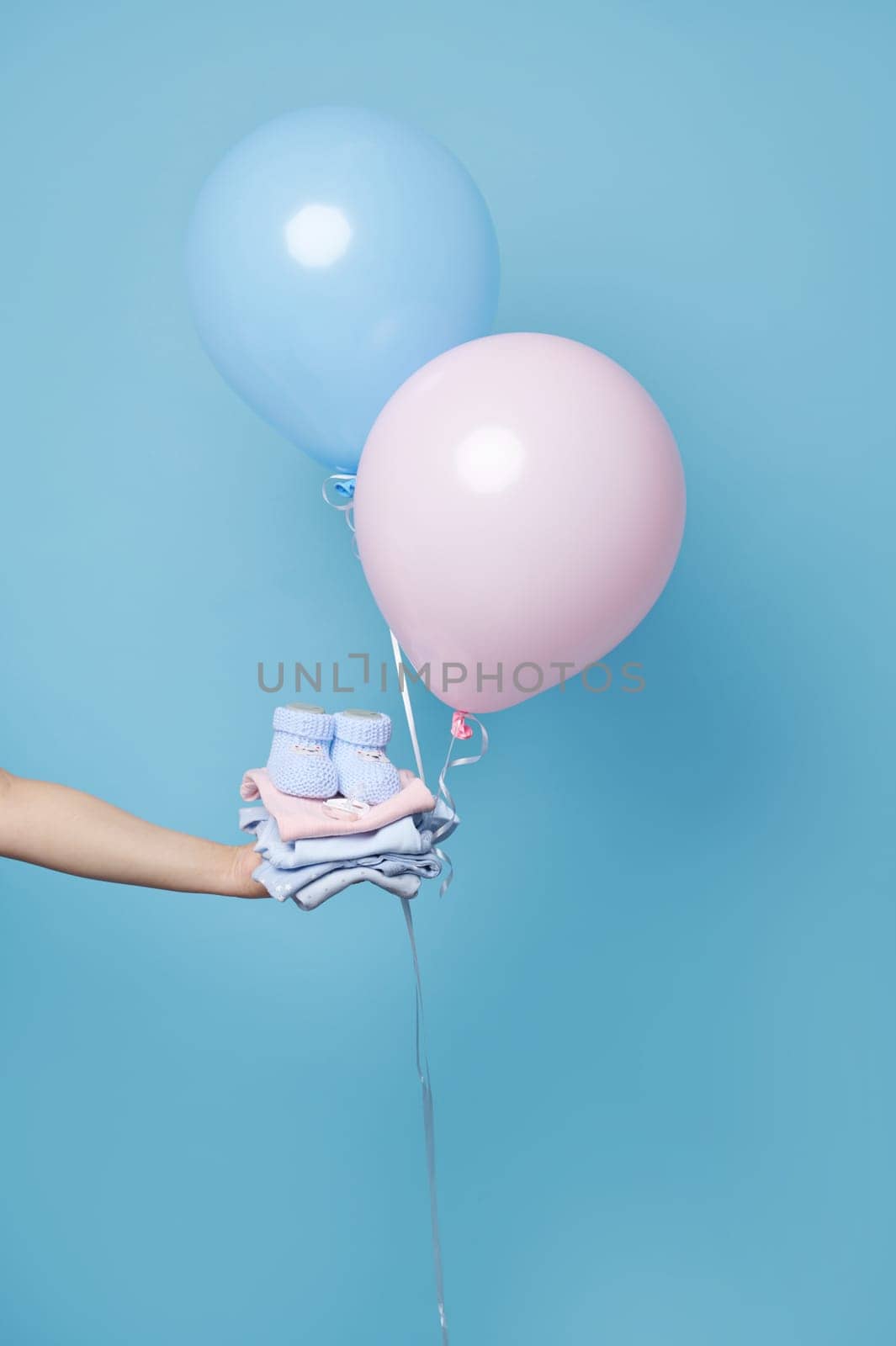 Hand holding stack of ironed laundered newborn baby clothes and booties, blue and pink balloons, isolated blue backdrop by artgf