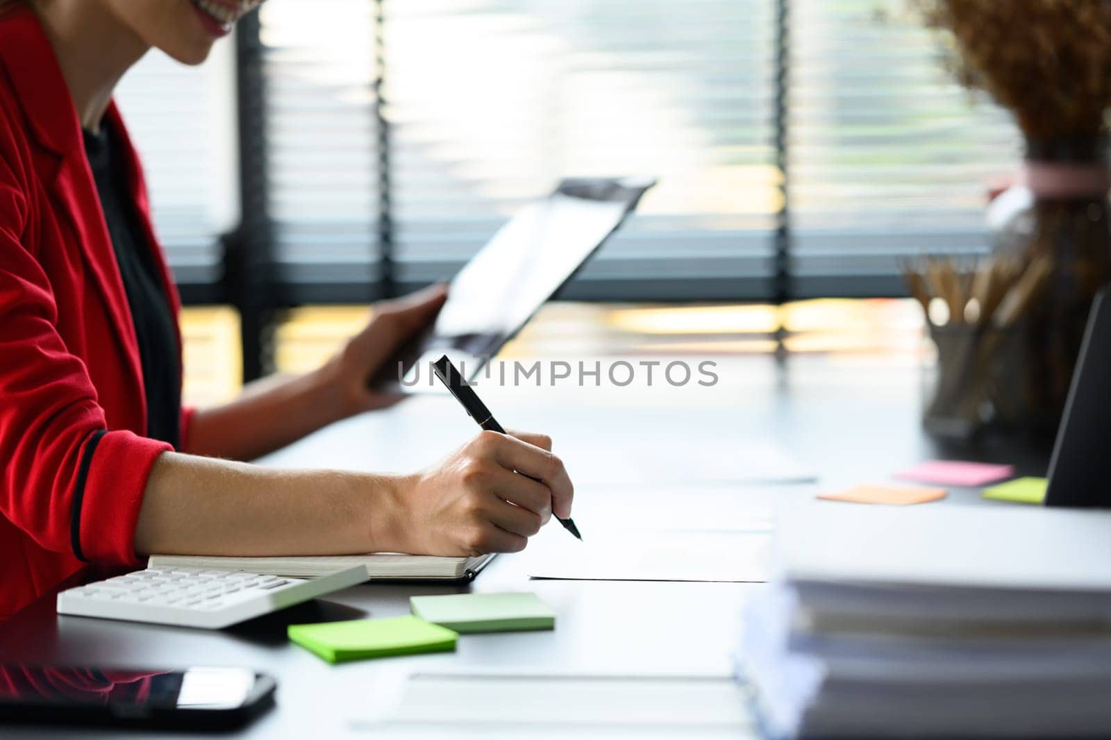 Side view of female entrepreneur holding digital tablet and writing important information on notepad.