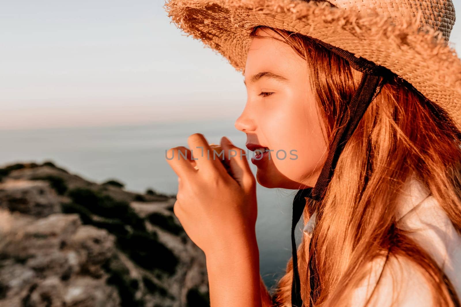 Happy girl eating corn. Summer snacking on the sea. Portrait of young beautiful girl in straw hat eating grilled corn while sitting by the sea on sunset time. Close up. Selective focus by panophotograph
