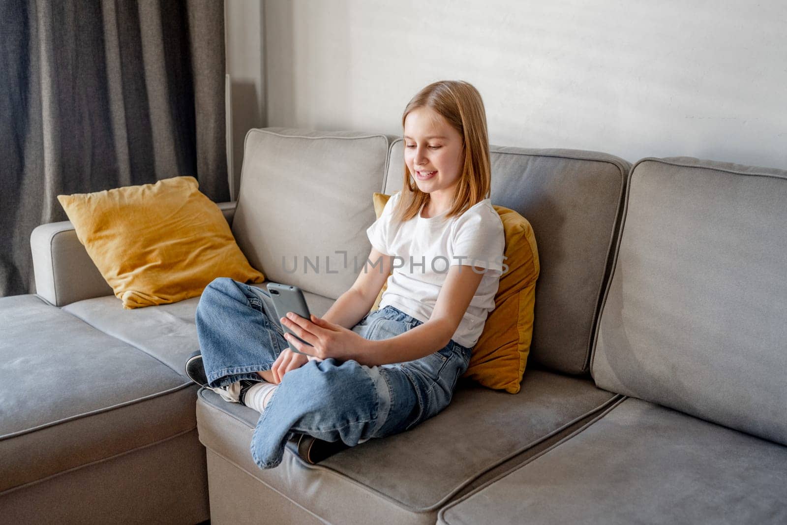 Preteen girl with smartphone sitting on sofa at home. Pretty child kid with modern mobile cellphone on couch