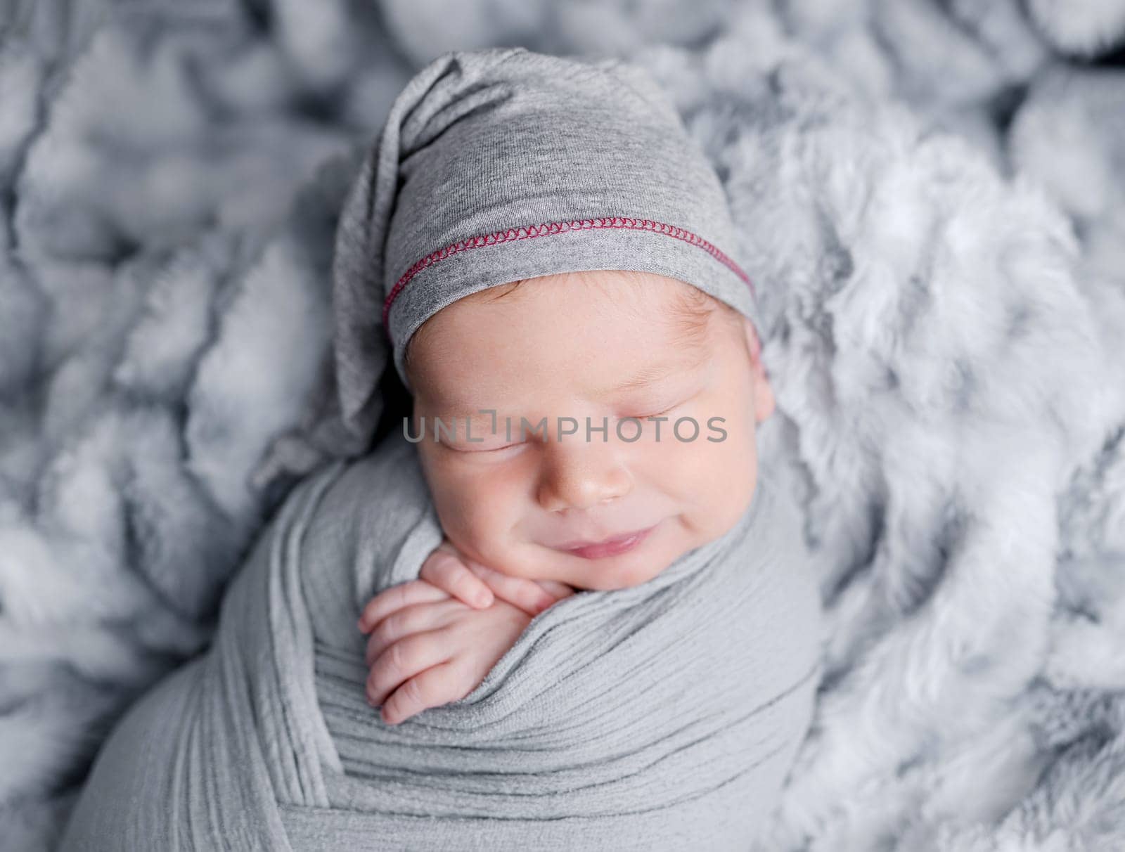 Cute newborn baby boy swaddled in grey fabric sleeping on fur blanket. Adorable infant child kid napping studio portrait