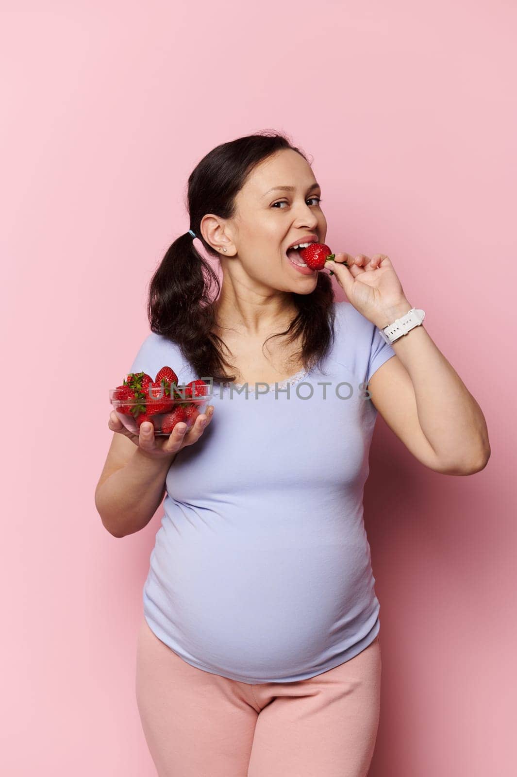 Charming glamour pregnant young woman with two ponytails, in blue t-shirt, looking at camera while eating fresh sweet strawberries out of a bowl, isolated on pink background. Pregnancy. Healthy eating