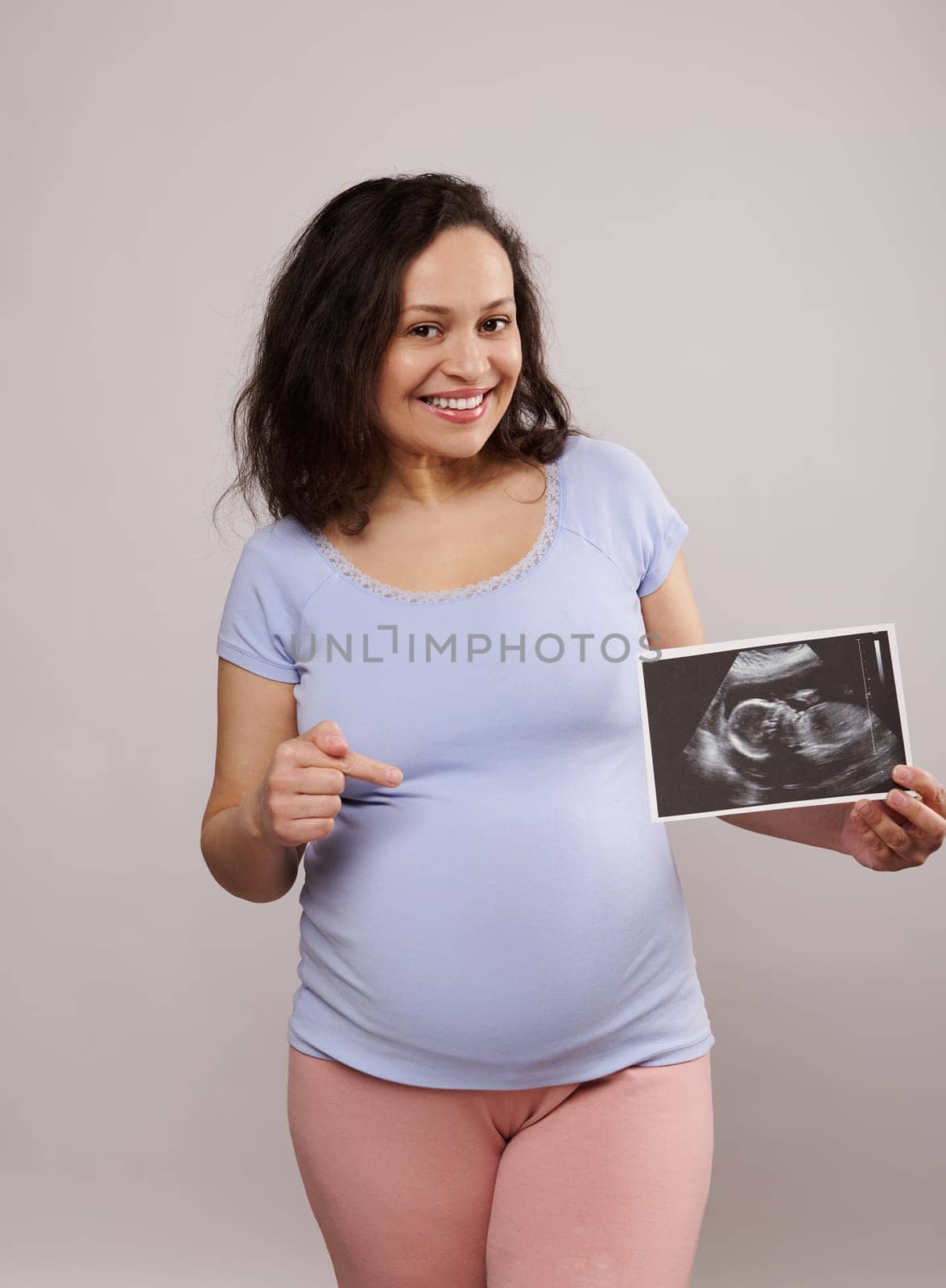 Smiling pregnant woman showing her newborn baby sonography to the camera and pointing at her belly, isolated on white by artgf