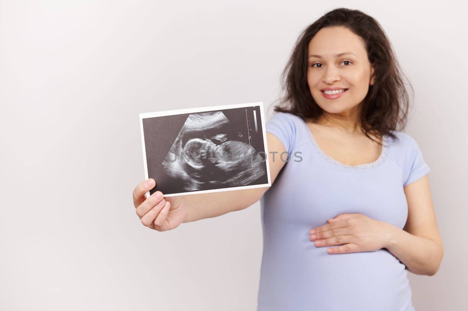 Selective focus on an ultrasound scan image, newborn baby sonography in the hand of a smiling charming pregnant woman, touching her belly, expressing positive emotions, isolated over white background