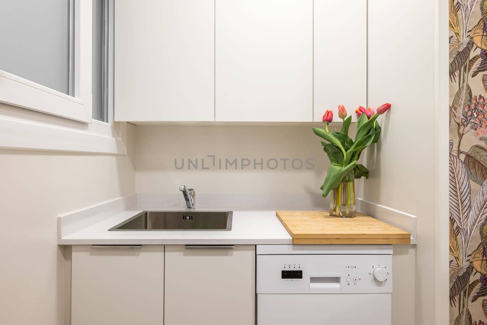 Close-up of a sink on a kitchen surface with cabinets and a vase of tulips in a modern stylish white kitchen. The concept of a stylish and comfortable kitchen in a small apartment. Copyspace.