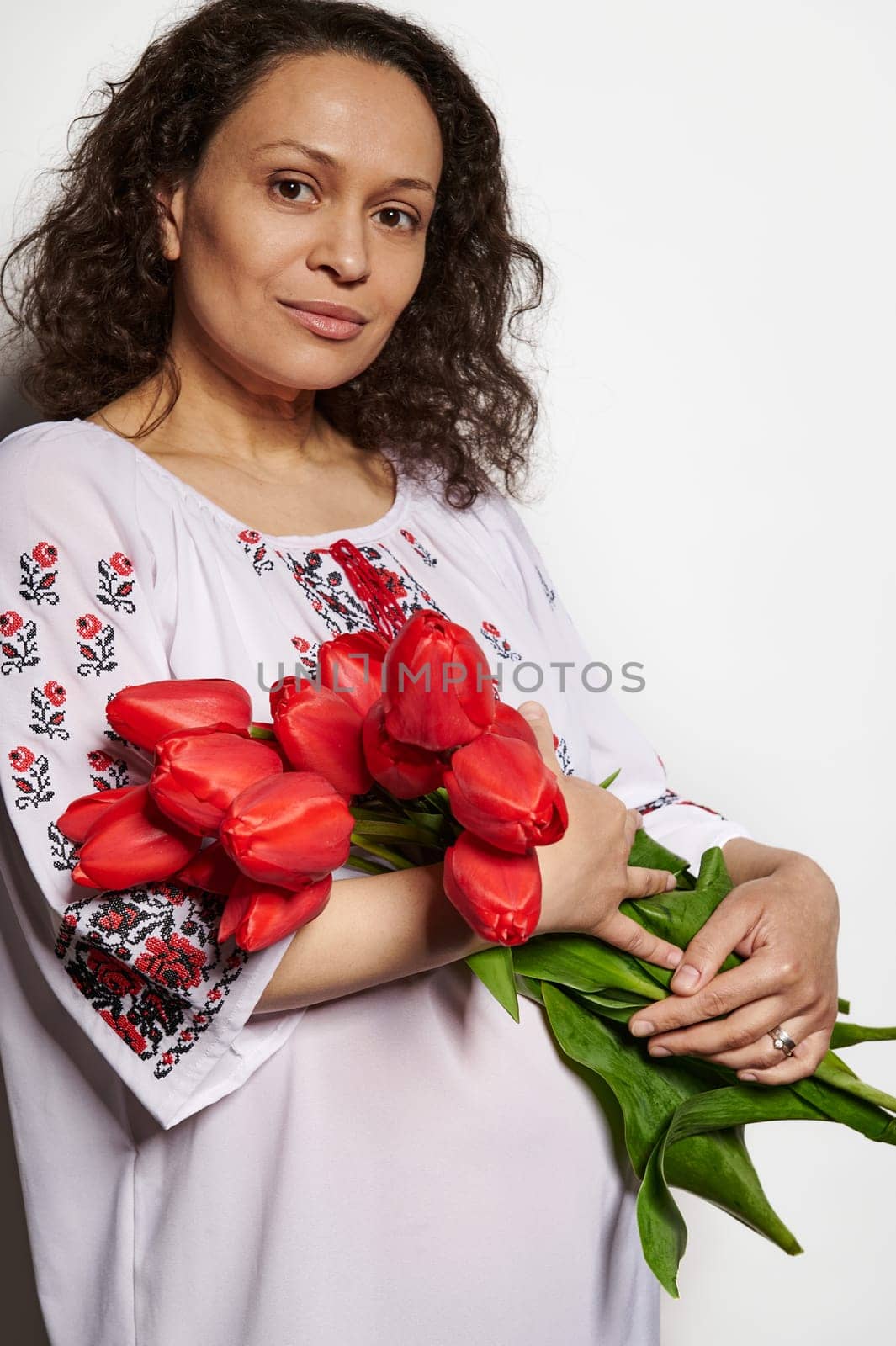 Confident portrait of a multi ethnic curly haired brunette, charming pregnant woman in traditional embroidered dress, holding a bouquet of red tulips, looking at camera over white isolated background.