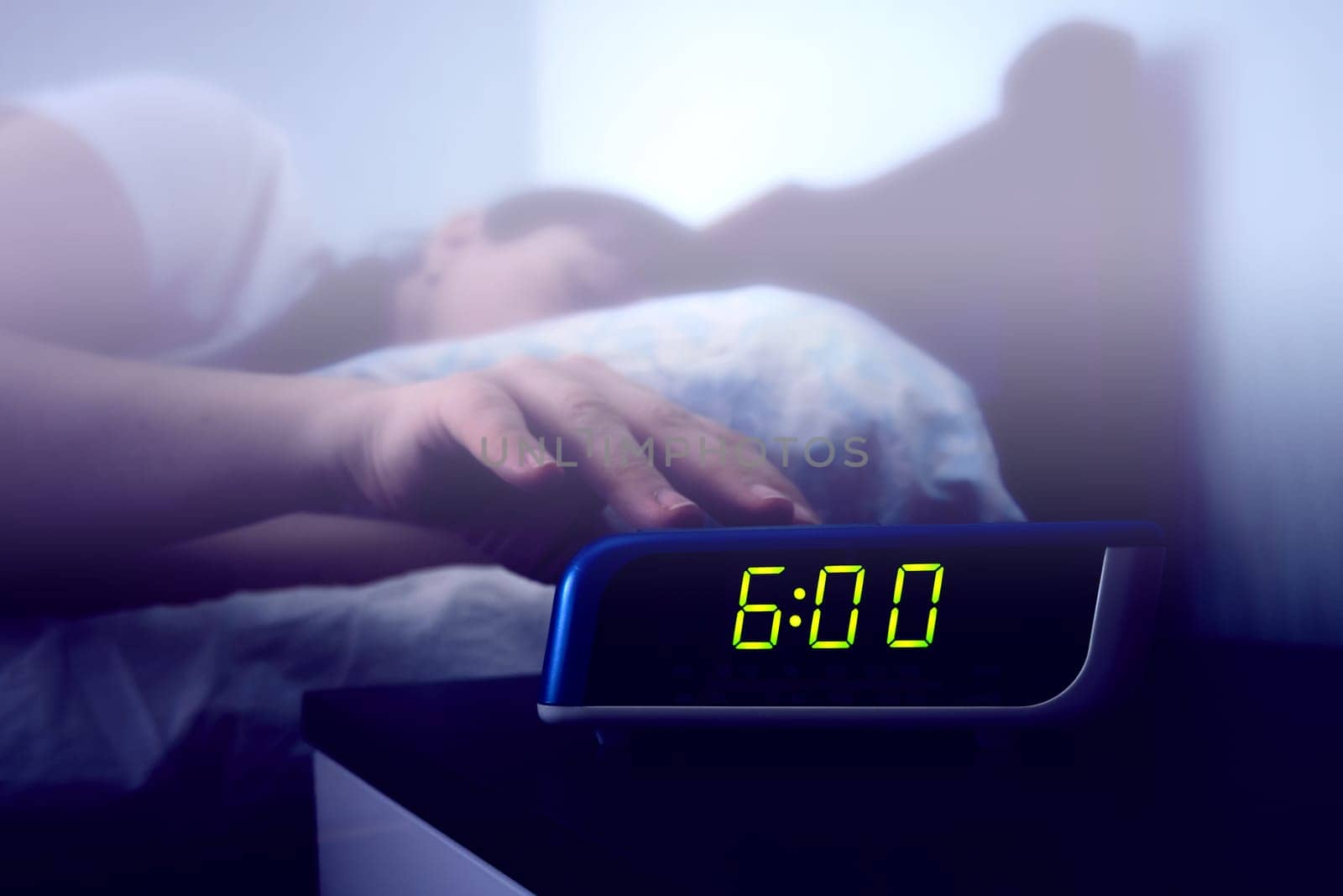 A sleeping woman's hand reaches for a digital alarm clock on the bedside table. Color toning and haze filter on the image